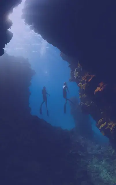 Two divers exploring an underwater cave with sunlight filtering through blue water